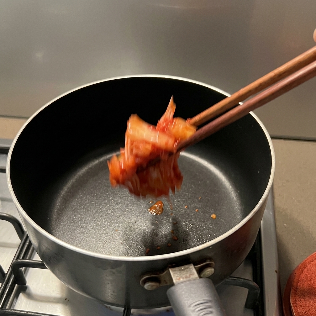 Rotes, gewürztes Kimchi wird mit Stäbchen in einen dunklen Topf auf dem Herd gegeben.
