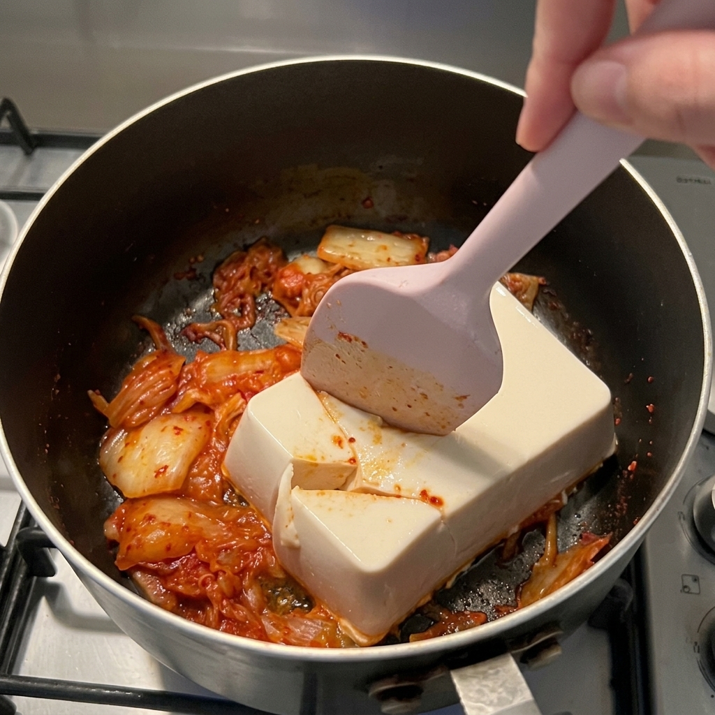 Ein rosa Silikonspatel teilt den weißen Seidentofu im Topf in grobe Stücke.