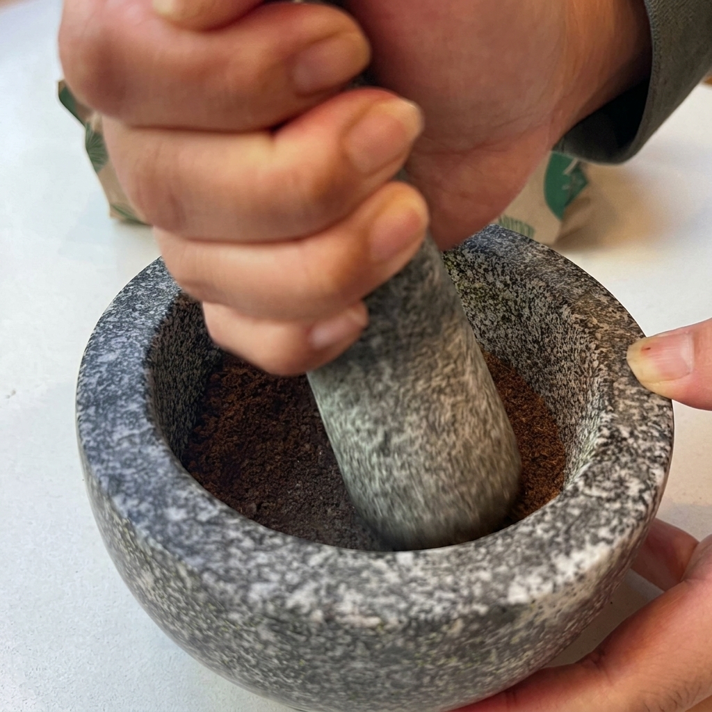 Hände mahlen geröstete Gewürze mit einem Stößel in einem Granitmörser zu einem feinen braunen Pulver.