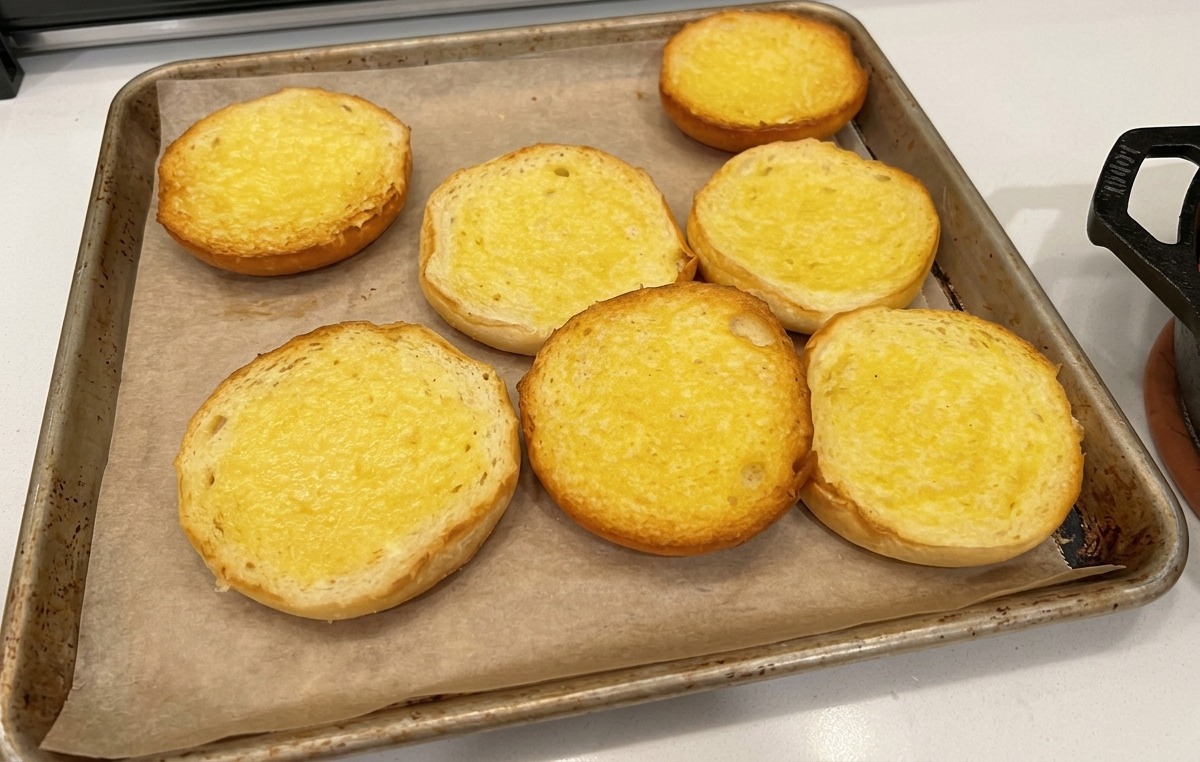 Butterbestrichene Hamburger-Brötchen liegen auf Backpapier auf einem Backblech.