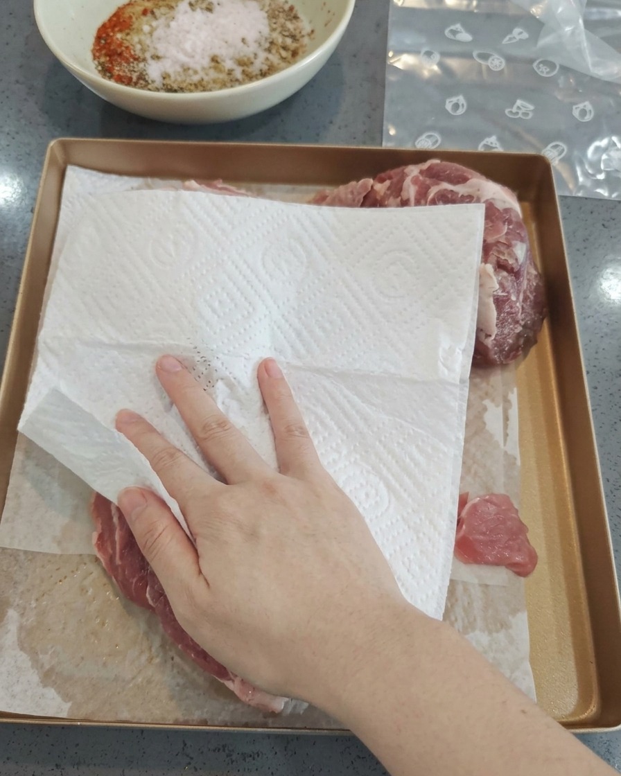 Eine Hand tupft mit einem weißen Küchentuch rohe Schweinefleischstücke auf einem Backblech trocken.