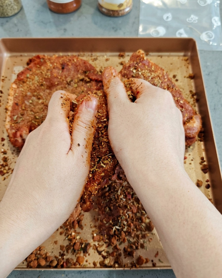 Hände reiben eine dunkle Gewürzmischung fest in große Stücke rohes Schweinefleisch ein.