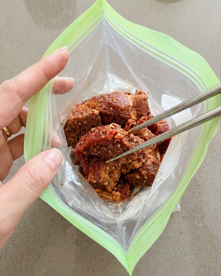 Gewürzte Schweinefleischstücke werden mit Stäbchen in einen grünen wiederverwendbaren Ziploc-Beutel gelegt.