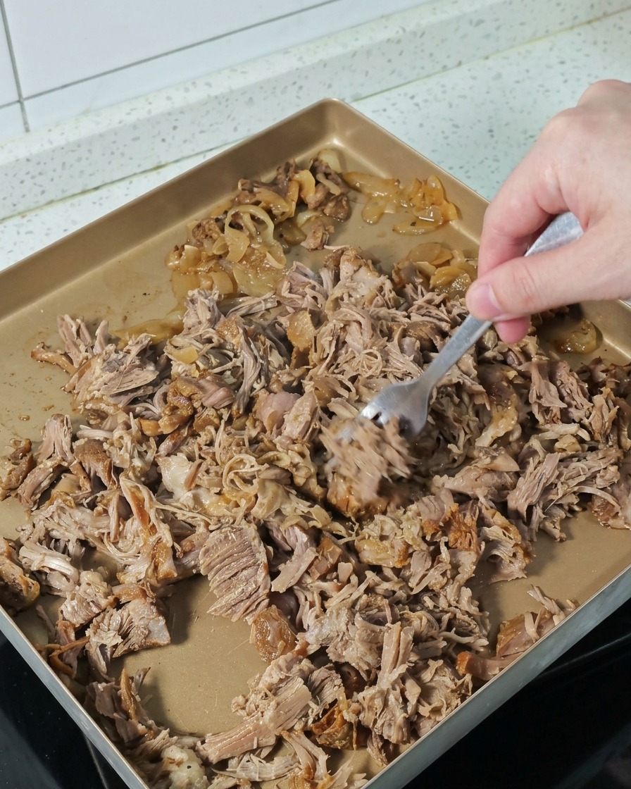 Eine Hand nutzt eine Gabel, um zartes Schweinefleisch auf einem Backblech in Stücke zu zupfen.
