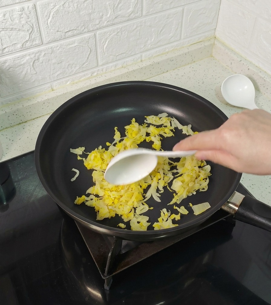 Eine Hand rührt gelbes Sauerkraut in einer schwarzen beschichteten Pfanne um.