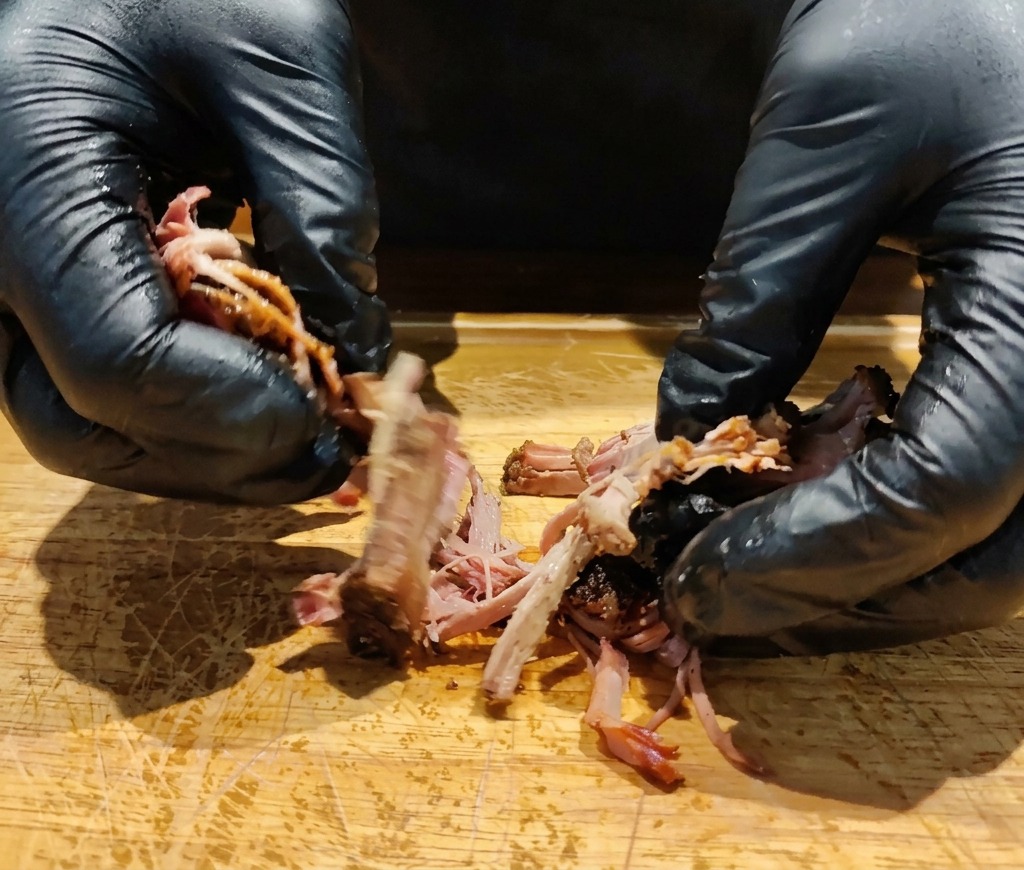 Hände mit Handschuhen zupfen ein Stück dunkel gerösteten Schweinenacken auf einem Holzbrett auseinander.