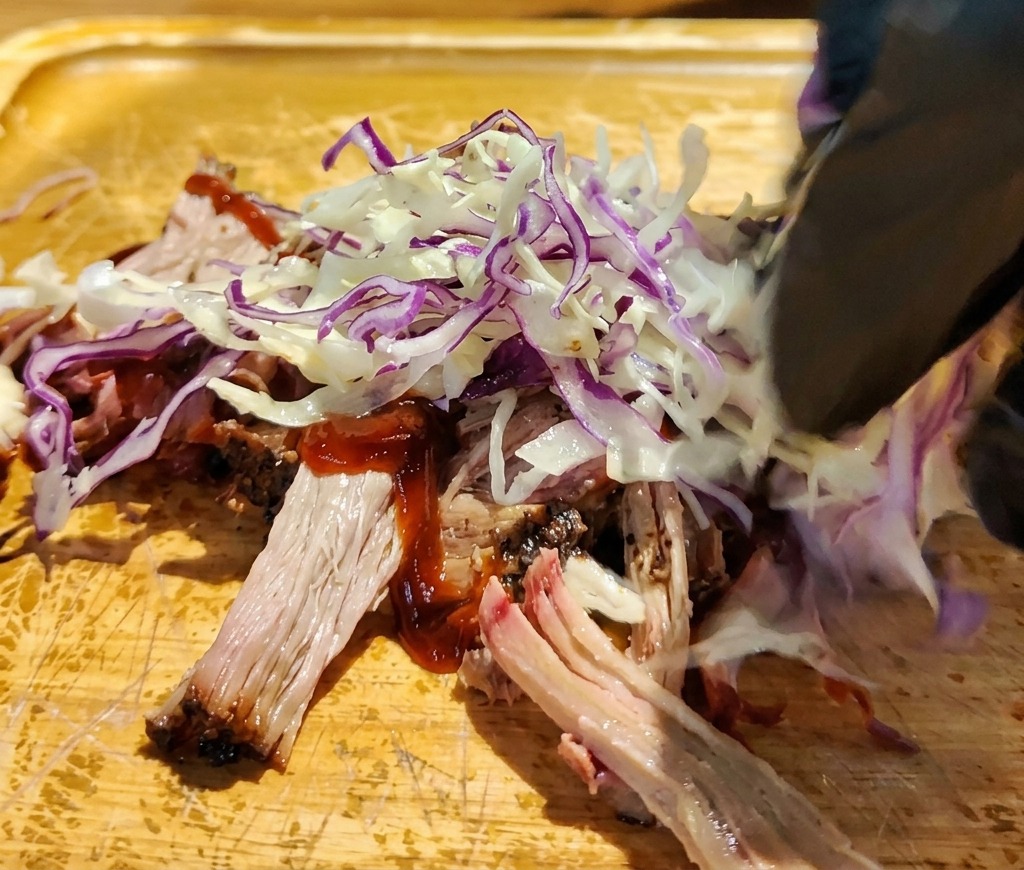 Eine Hand legt frischen Rot- und Weißkohlsalat auf das marinierte Pulled Pork.