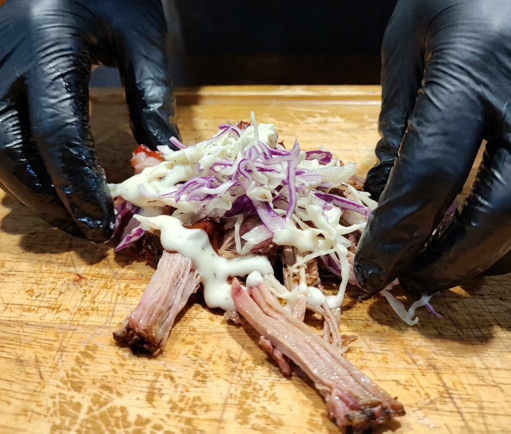Hände neben einem Haufen Pulled Pork, der mit Krautsalat und weißem cremigen Dressing getoppt ist.