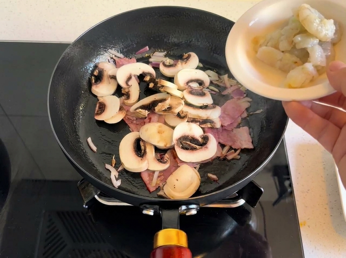 Rohe graue Garnelen werden in eine Pfanne zu angebratenem Speck, Pilzen und Zwiebeln gegeben.