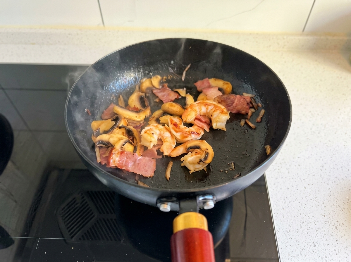 Garnelen, Speck und Pilze werden in einer schwarzen Pfanne gebraten, bis die Garnelen komplett rosa sind.