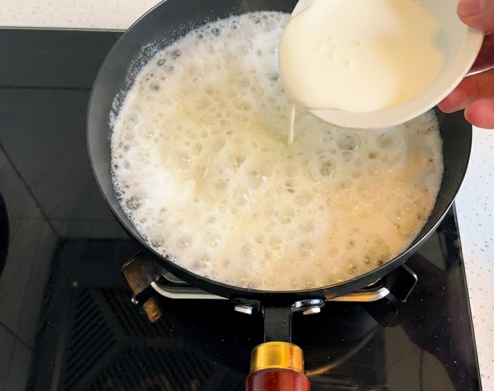 Dickflüssige Schlagsahne wird aus einer Schale in die köchelnde helle Suppenbasis gegossen.