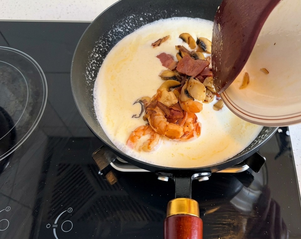 Ein Teller mit gebratenen Garnelen, Speck und Pilzen wird in die cremige Suppenbasis in der Pfanne geleert.