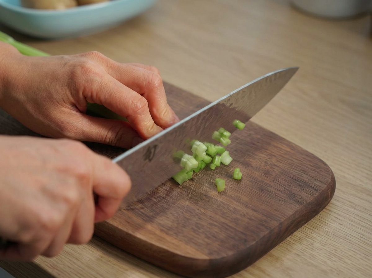 Ein Kochmesser schneidet leuchtend grüne Staudensellerie-Stangen auf einem Holzbrett.