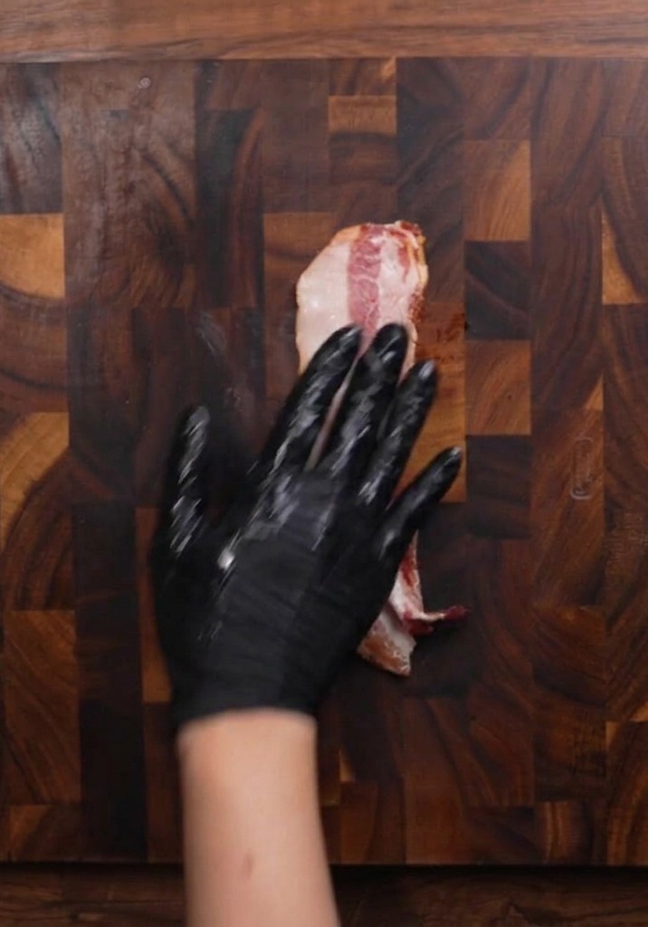 Eine Hand mit Handschuh hält ein Stück rohen Speck auf einem dunklen Holzbrett fest, um ihn zu schneiden.
