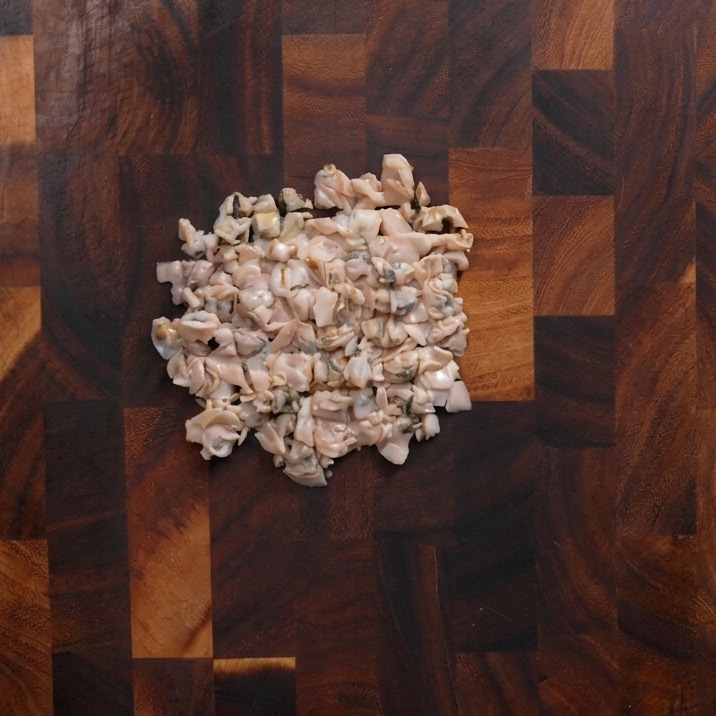 Ein Haufen grob gehacktes Muschelfleisch liegt in der Mitte eines dunklen Holzschneidebretts.