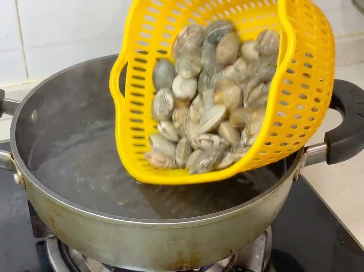 Ganze Muscheln werden aus einem gelben Sieb in einen Topf mit kochendem Wasser geschüttet.