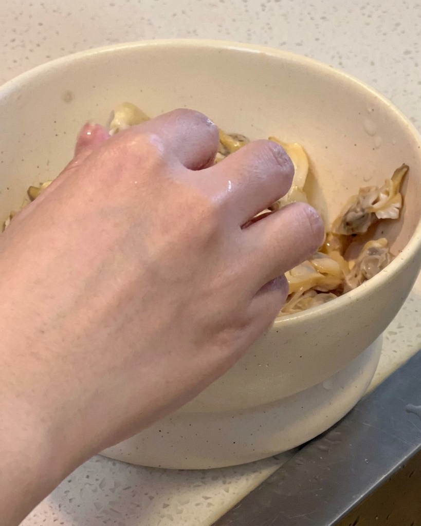 Eine Hand löst das gegarte Muschelfleisch aus der Schale und gibt es in eine weiße Schüssel.