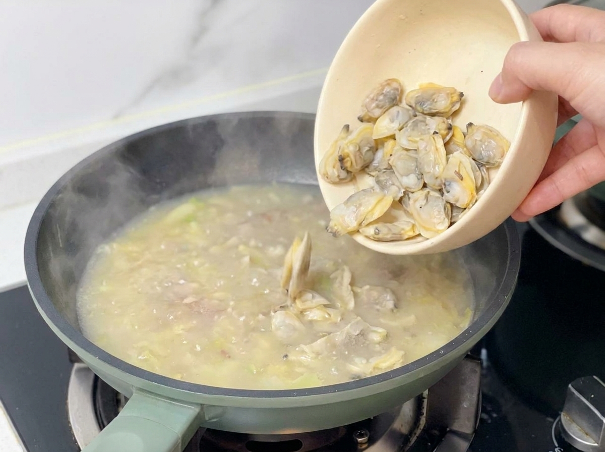 Gegartes Muschelfleisch wird aus einer beigen Schüssel in die köchelnde Suppenbasis gegeben.