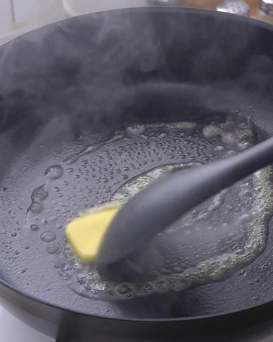 Ein Stück gelbe Butter schmilzt in einer heißen schwarzen Pfanne und wird mit einem Pfannenwender bewegt.