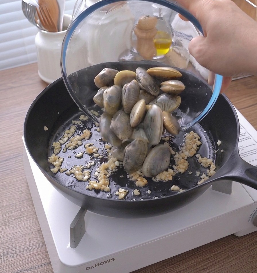 Ganze Venusmuscheln werden aus einer Glasschüssel in die Pfanne zu Knoblauch und Butter geschüttet.