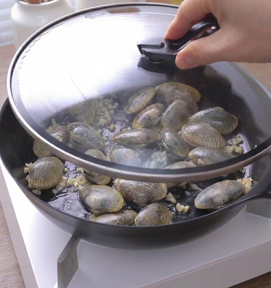 Eine Hand setzt einen Glasdeckel auf die Pfanne mit Muscheln und Knoblauch.