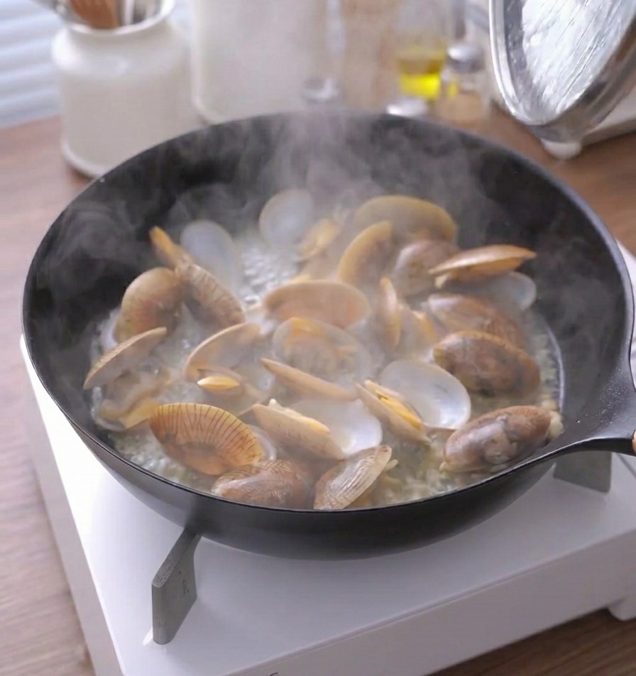 Dampf steigt aus der Pfanne mit geöffneten, gegarten Venusmuscheln auf.