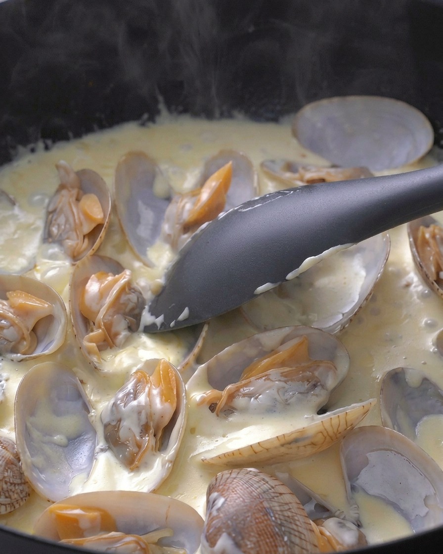 Ein Pfannenwender rührt die Muscheln vorsichtig in der köchelnden weißen Sahnesauce um.