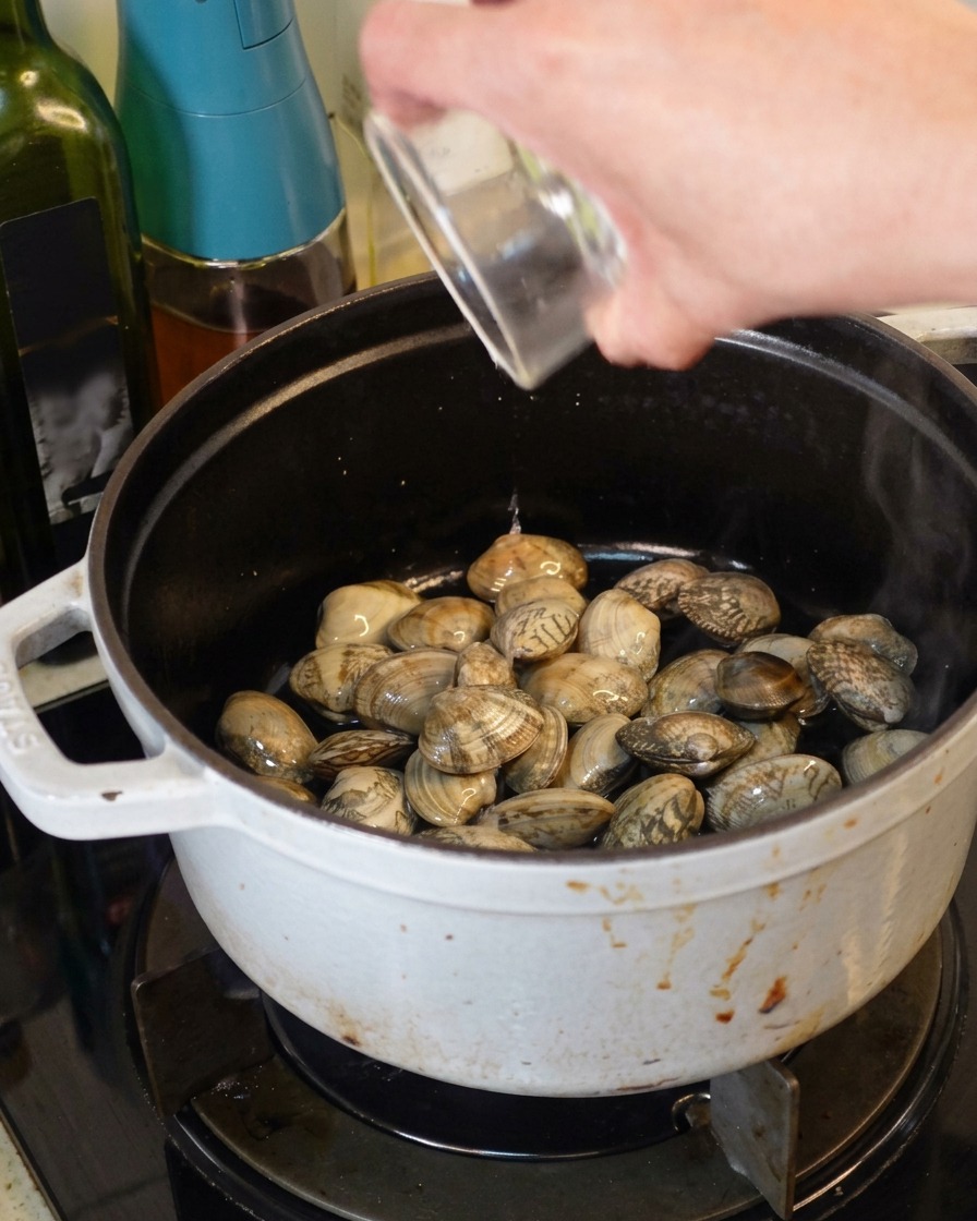 Weißwein wird aus einem Glasbehälter in einen weißen gusseisernen Topf mit frischen Muscheln gegossen.