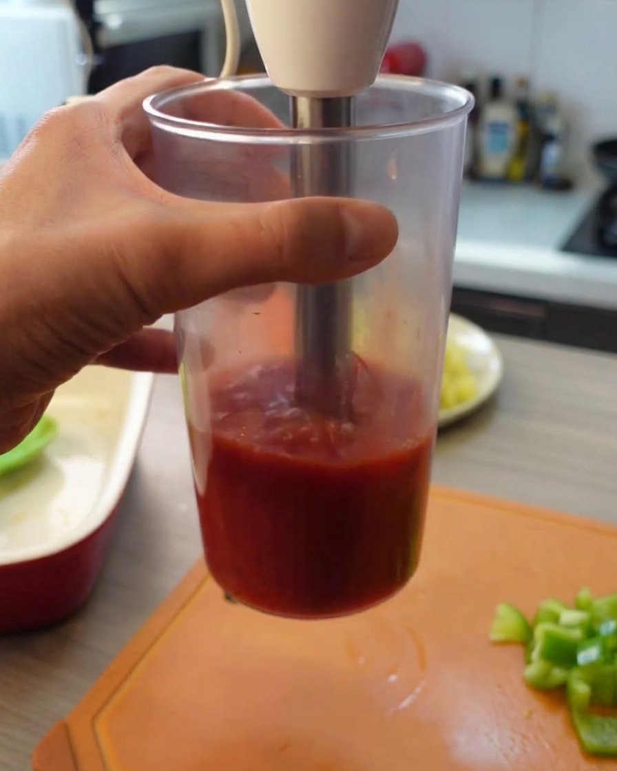 Ein Stabmixer püriert Dosentomaten in einem hohen Kunststoffbecher.