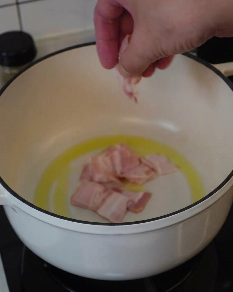 Eine Hand gibt Speckstücke in einen weißen Topf mit etwas Öl zum Anbraten.