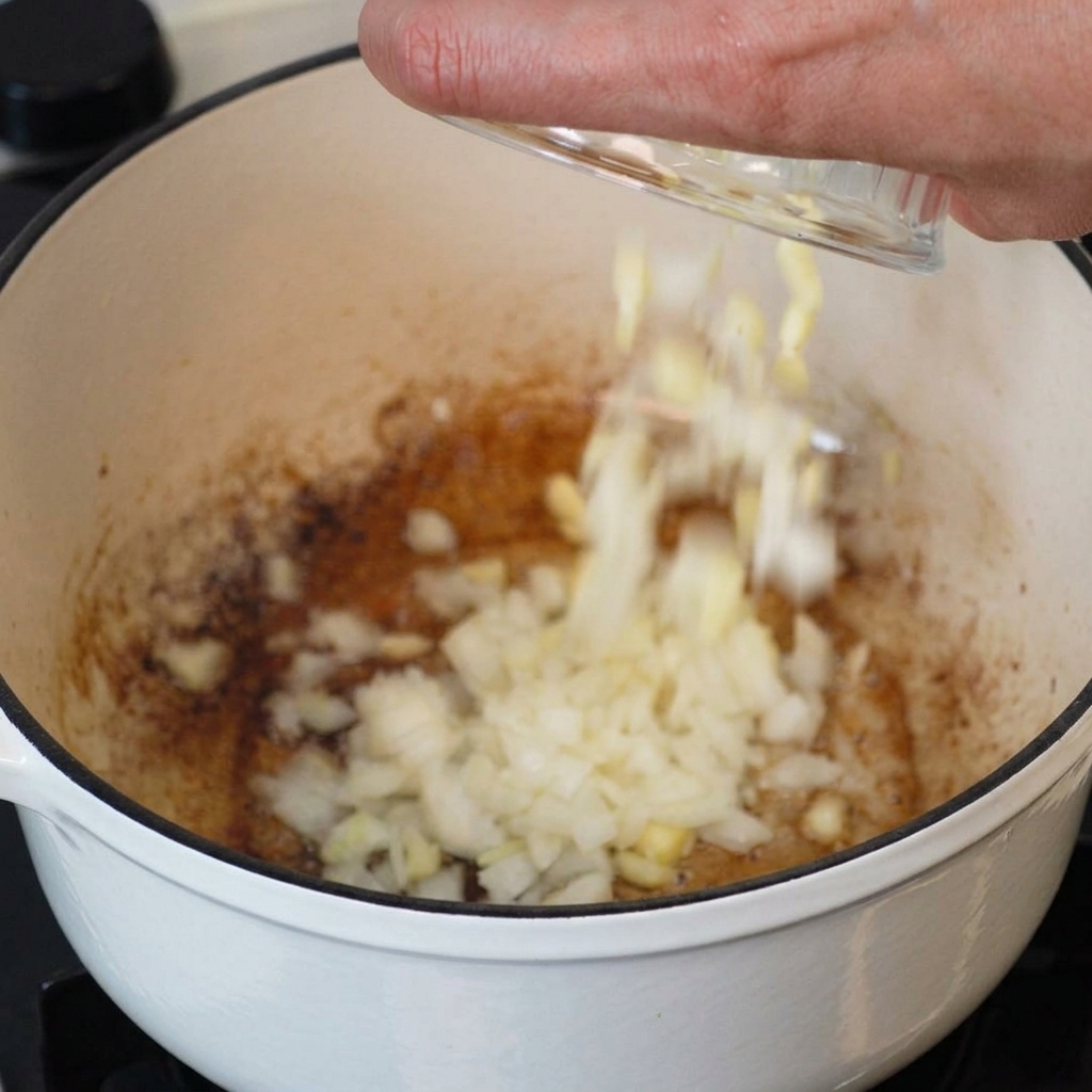Eine Hand gibt Zwiebeln und Knoblauch in den Topf mit dem verbliebenen Speckfett.
