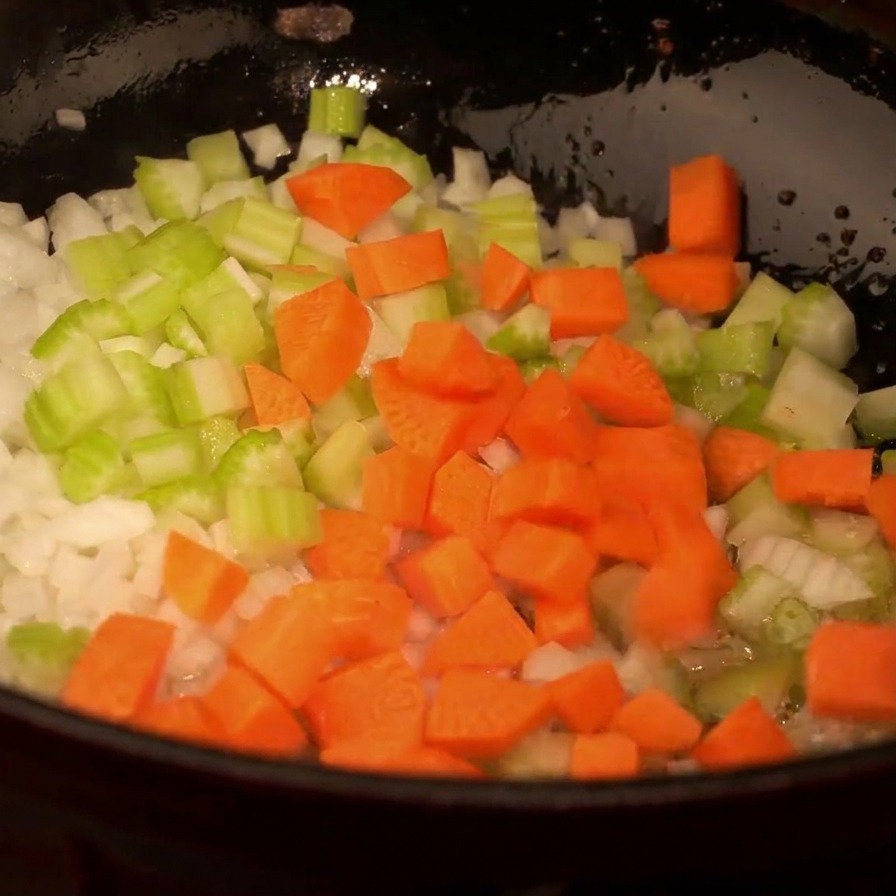 Draufsicht auf eine Pfanne mit Zwiebeln, Sellerie und leuchtend orangen Karottenwürfeln.