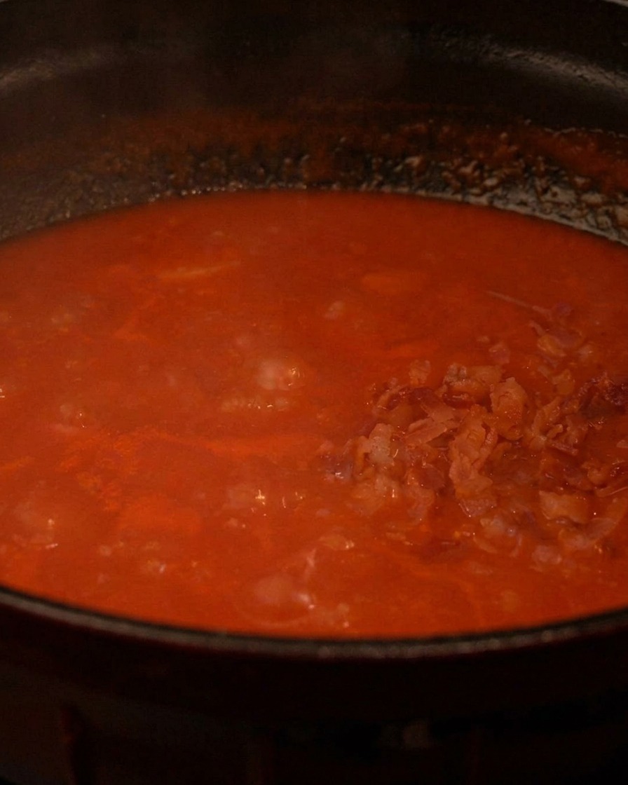 Knusprige Speckwürfel werden in einen Topf mit dampfender roter Suppe gegeben.