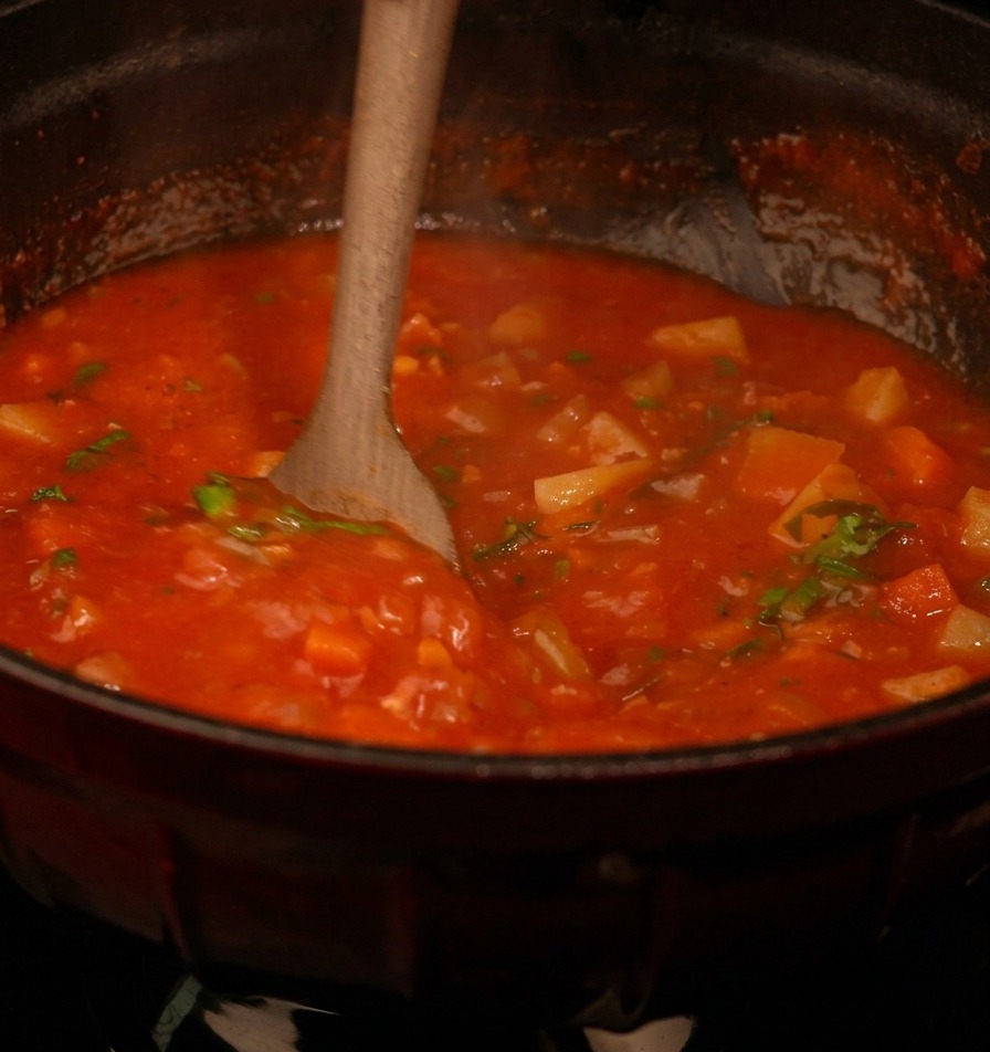Ein Holzlöffel rührt die rote Suppe mit Gemüsestücken und grüner Petersilie um.