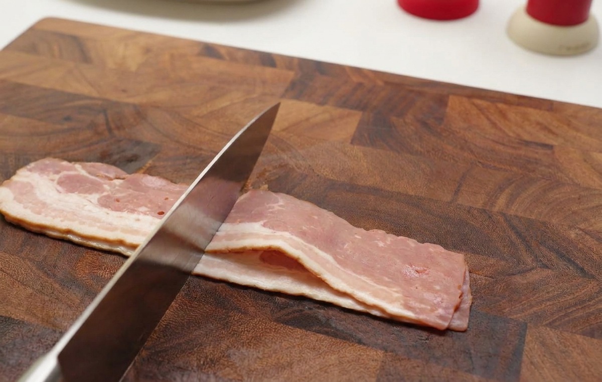 Hände schneiden rohe Speckstreifen mit einem scharfen Messer auf einem Holzbrett in kleine Stücke.