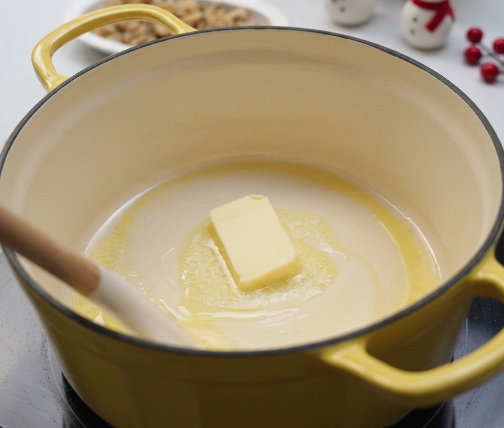 Ein Stück Butter schmilzt in einem gelben Topf bei niedriger Hitze und wird mit einem Teigschaber gerührt.