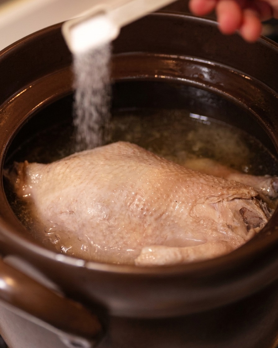 Eine Hand streut mit einem kleinen weißen Messlöffel Salz über ein ganzes, gekochtes Huhn in klarer Brühe.