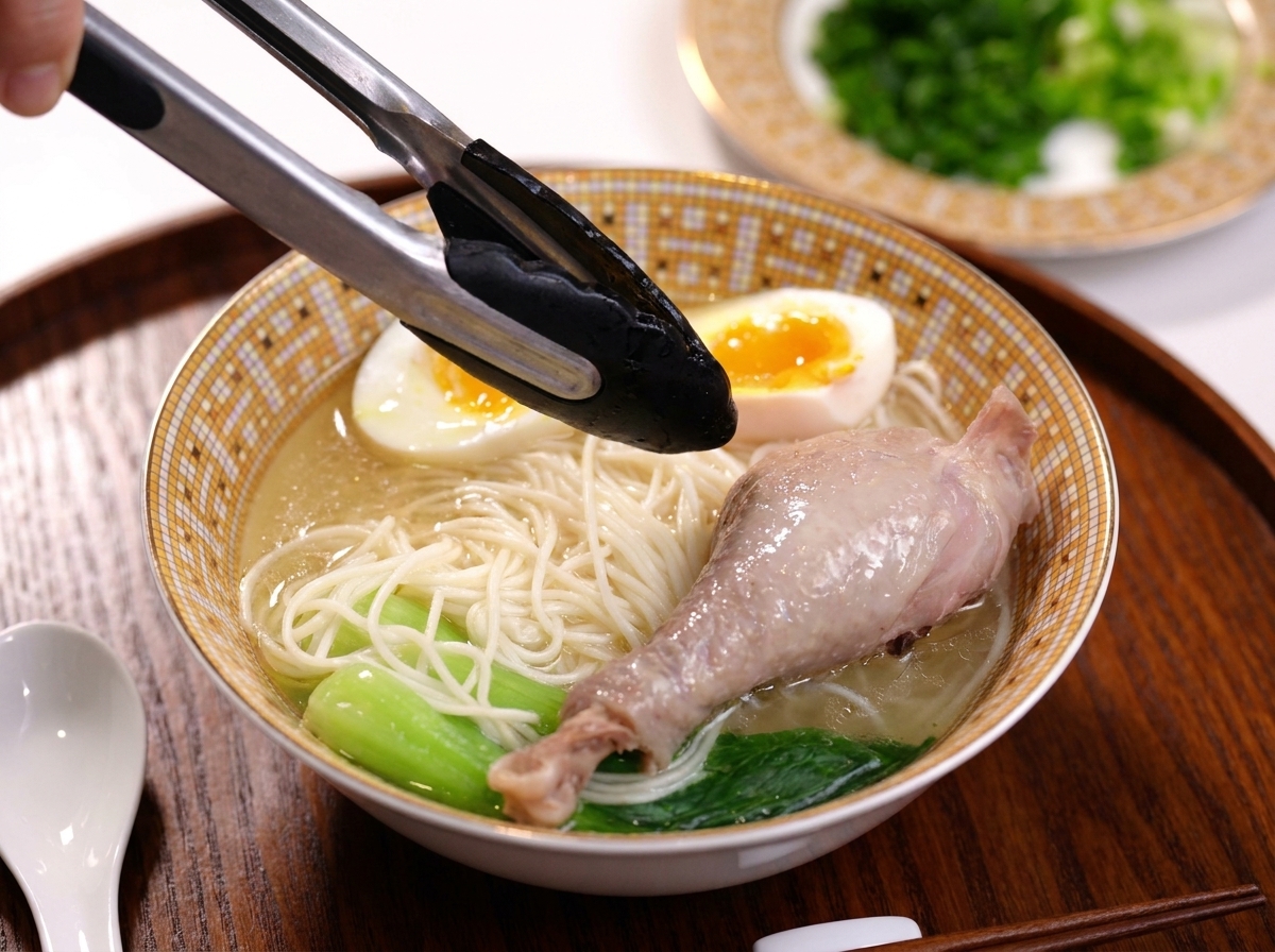 Eine Zange legt eine gekochte Hähnchenkeule in eine Schüssel mit Hühnersuppe, Pak Choi und einem weichgekochten Ei.
