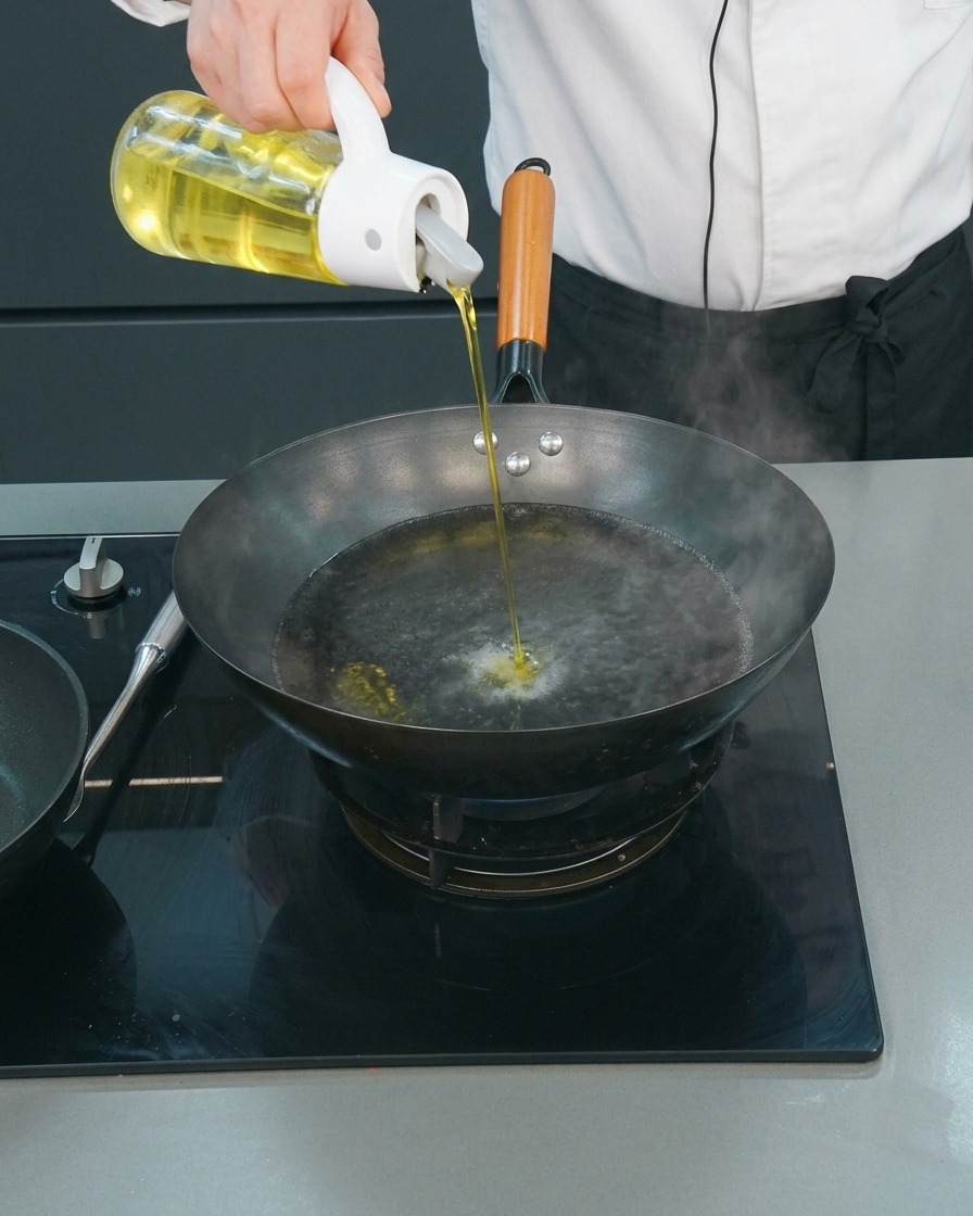 Ein Koch gießt aus einem Glasspender einen dünnen Strahl Speiseöl in einen Wok mit kochendem Wasser.