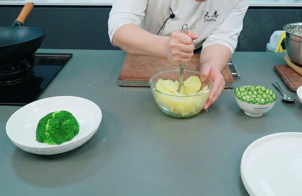Ein Koch zerdrückt gedämpfte Kartoffeln in einer klaren Glasschüssel auf einer Arbeitsplatte mit einem Kartoffelstampfer aus Metall.