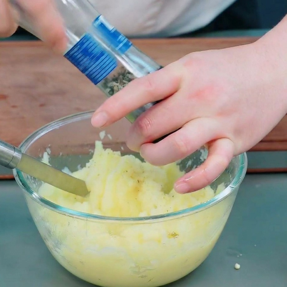 Hände würzen feines Kartoffelpüree in einer klaren Glasschüssel mit einer Salzmühle und frisch gemahlenem schwarzen Pfeffer.