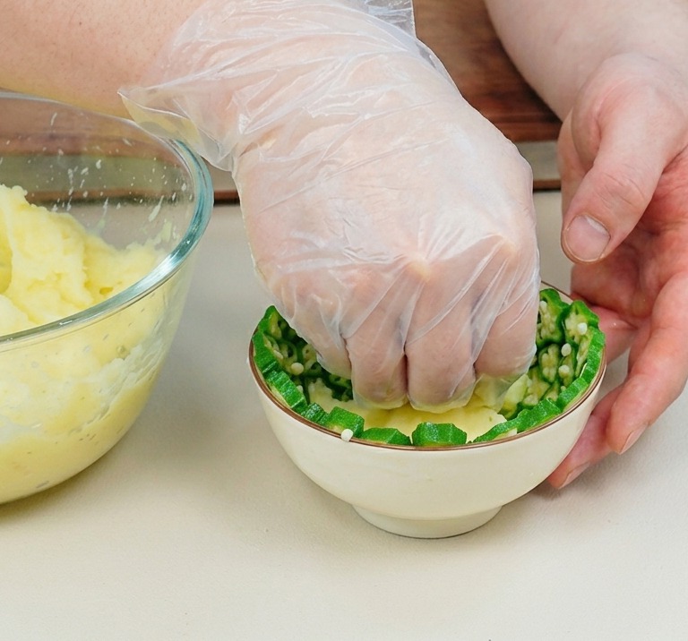 Eine Hand in einem Plastikhandschuh drückt gelbes Kartoffelpüree in eine kleine weiße Schüssel, die mit grünen, sternförmigen Okrascheiben ausgekleidet ist.