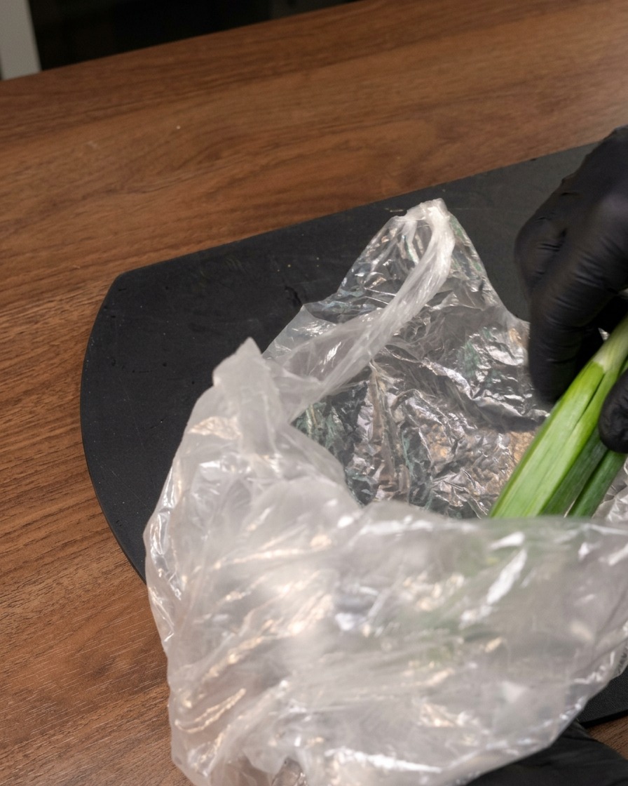 Eine behandschuhte Hand nimmt frische grüne Okraschoten aus einer Plastiktüte auf einer dunklen Oberfläche.