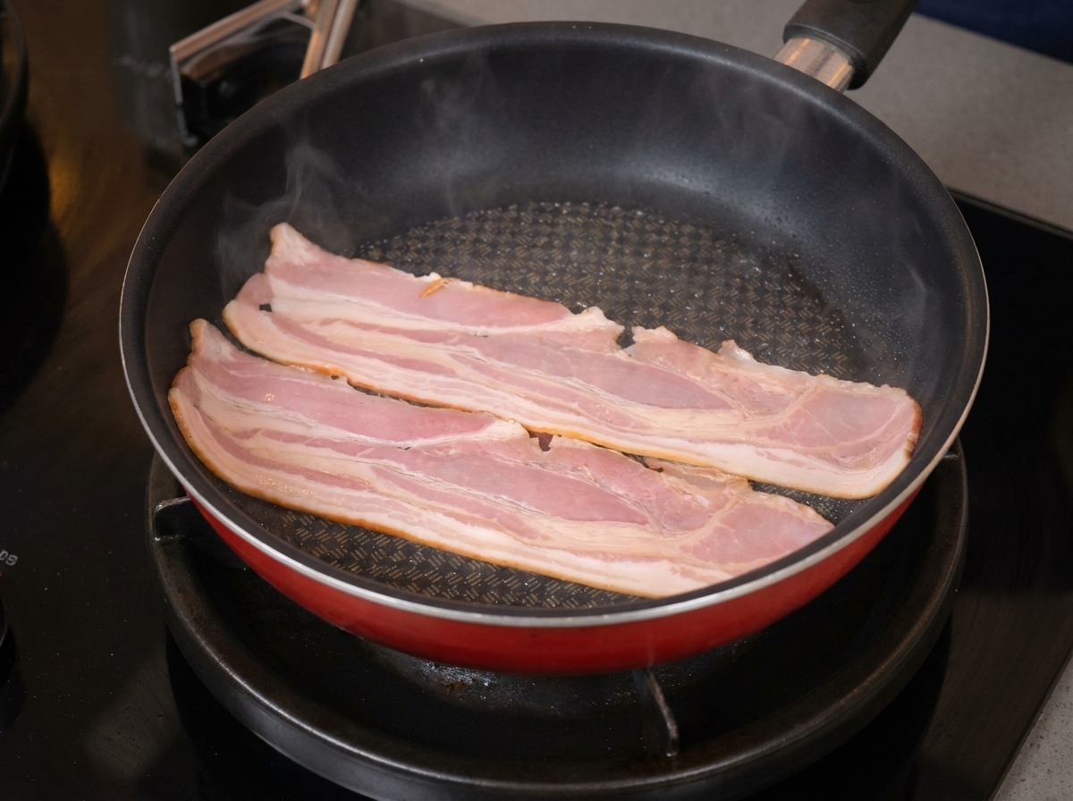 Zwei lange Streifen Speck brutzeln in einer roten beschichteten Pfanne und beginnen sich zu wellen.
