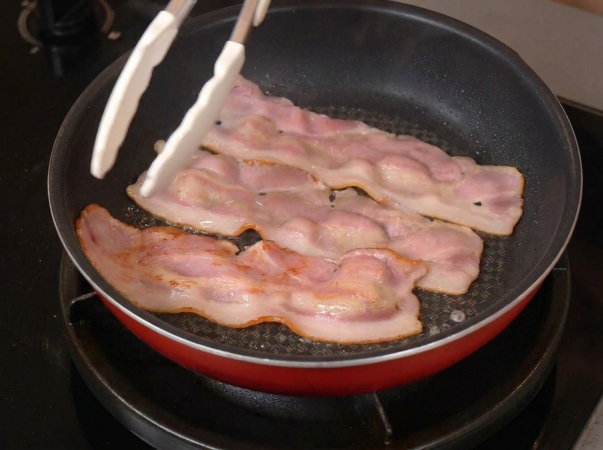 Drei brutzelnde Speckstreifen in einer Pfanne, die mit einer weißen Zange gewendet werden.