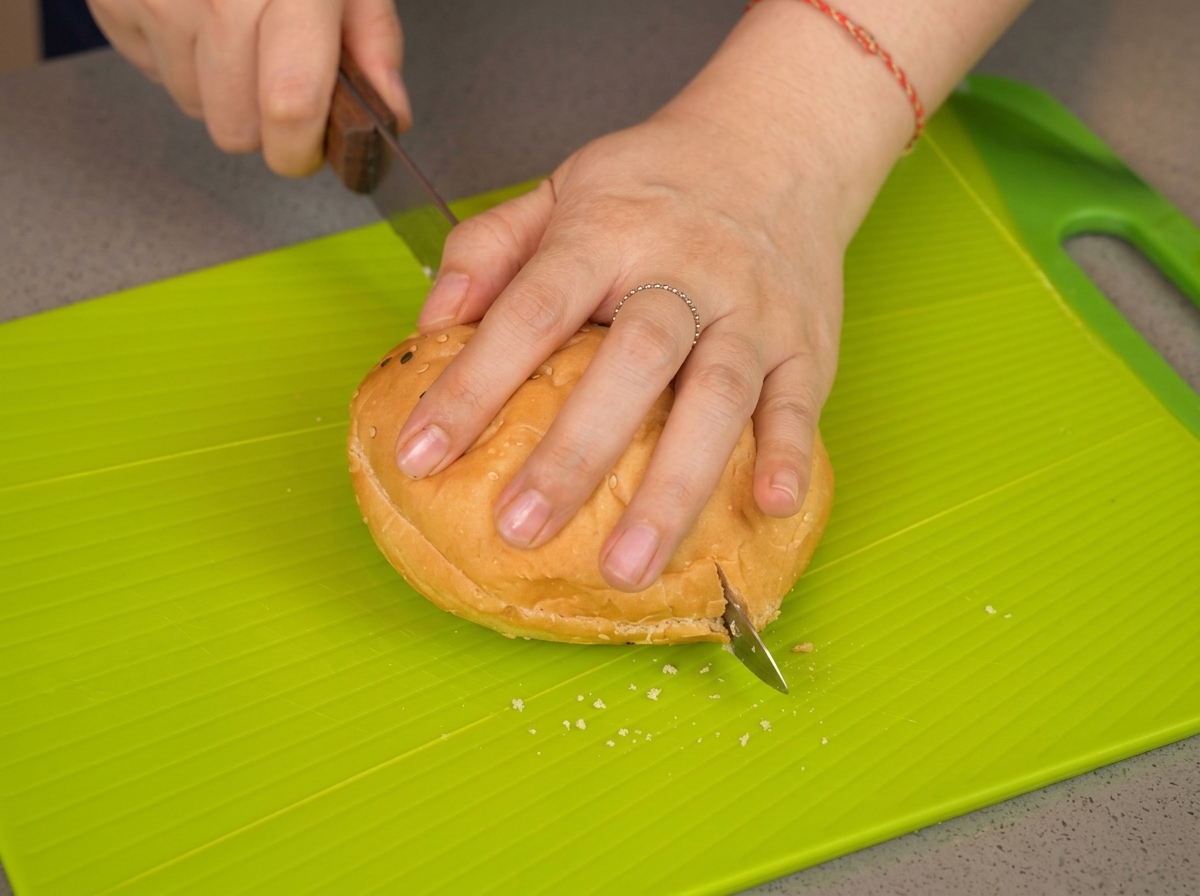 Eine Hand drückt ein Sesambrötchen auf einem hellgrünen Schneidebrett nach unten, während ein Messer es in der Mitte durchschneidet.