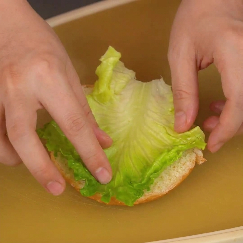 Hände legen ein leuchtend grünes Salatblatt auf die Schnittfläche der unteren Brötchenhälfte.