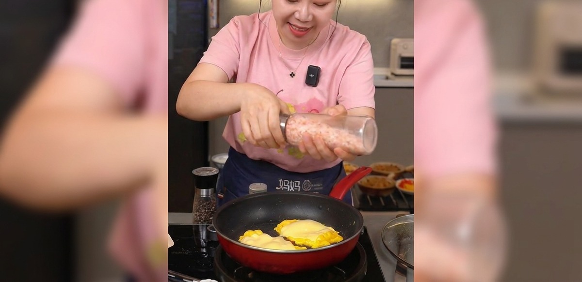 Hände mahlen rosa Meersalz aus einer Mühle über eine Pfanne mit fertigem Rührei und geschmolzenem Käse.