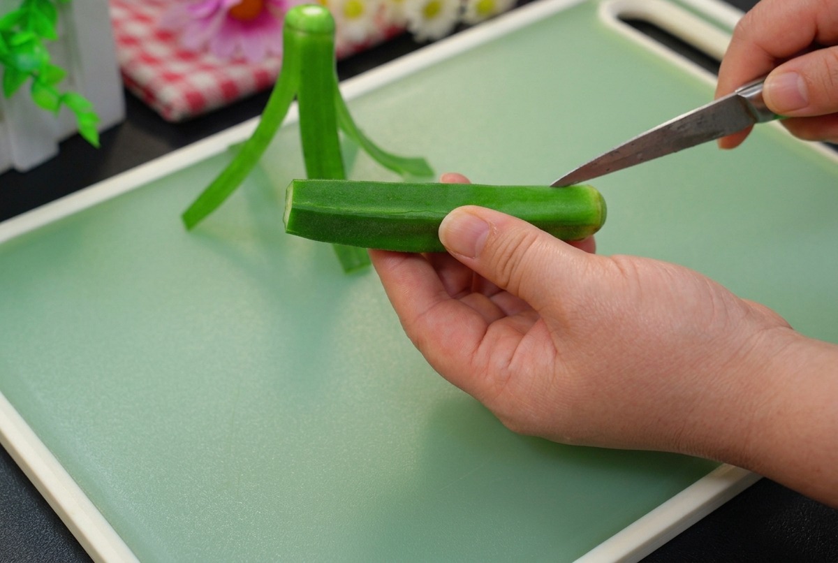Abschneiden der Spitze einer frischen grünen Okraschote mit einem kleinen Gemüsemesser auf einem hellgrünen Schneidebrett.