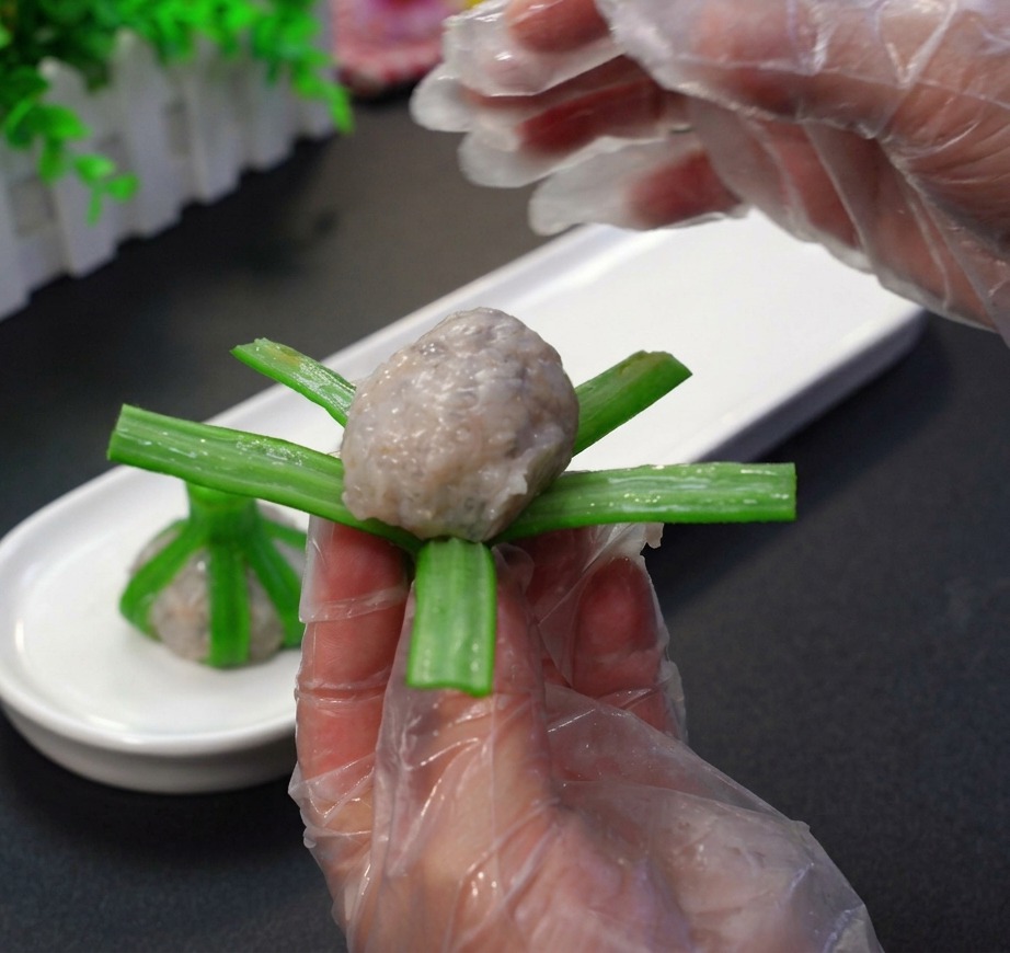 Hände mit Handschuhen, die rohe Garnelenpaste zu einer Kugel formen und diese in die grünen Streifen einer blanchierten Okraschote einwickeln.
